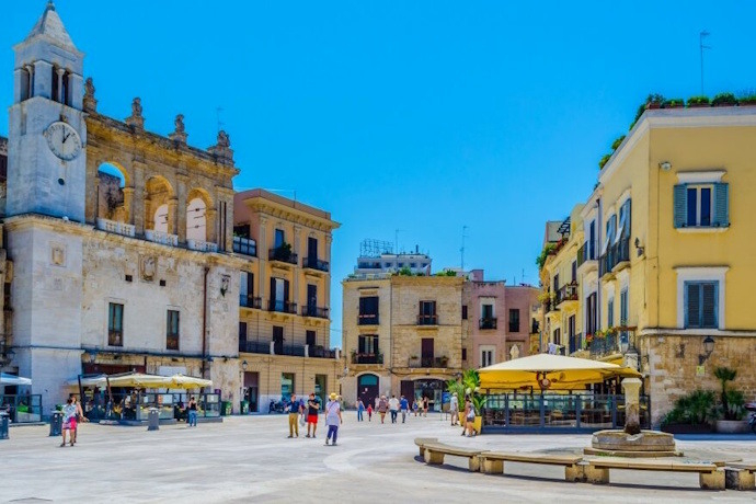 A sunny plaza in Bari, Italy, surrounded by historic buildings, outdoor cafés, and people enjoying the open space.