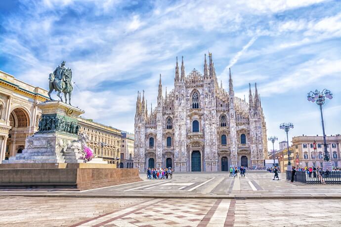 The majestic façade of the Milan Cathedral (Duomo di Milano) in Piazza del Duomo, with people walking across the square and a statue of King Victor Emmanuel II on horseback in the foreground.