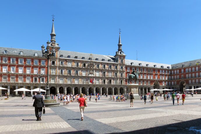 Wide view of Madrid’s Plaza Mayor, with red-brick arcades, a central equestrian statue, and crowds crossing the sunny square.