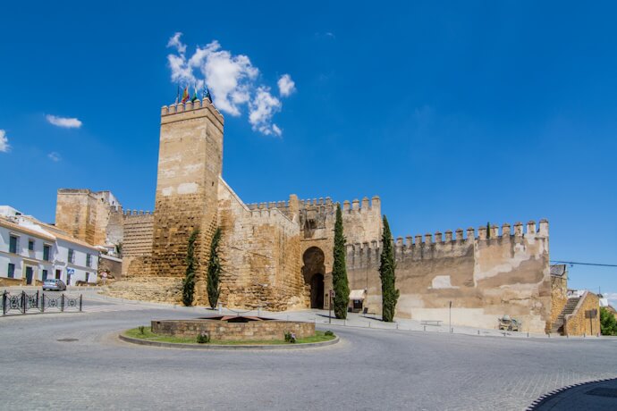 Medieval fortress walls and gate in Carmona, built from golden stone and set atop a hill under a clear blue sky.