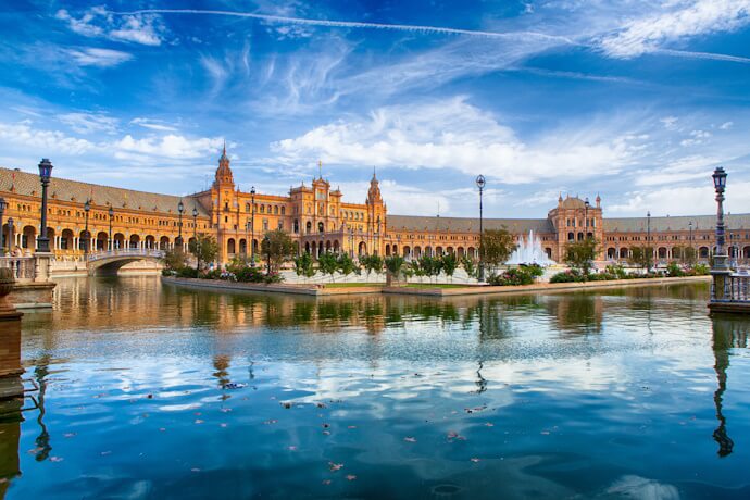 Plaza de España in Seville reflected in the canal, with ornate towers, bridges, and warm-toned architecture under a bright blue sky.