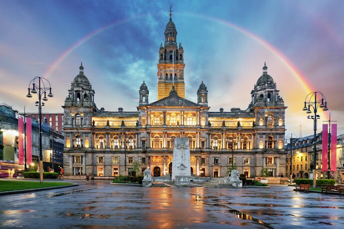 The illuminated façade of Glasgow’s City Chambers at dusk, with a glowing rainbow arching across the sky above the square.