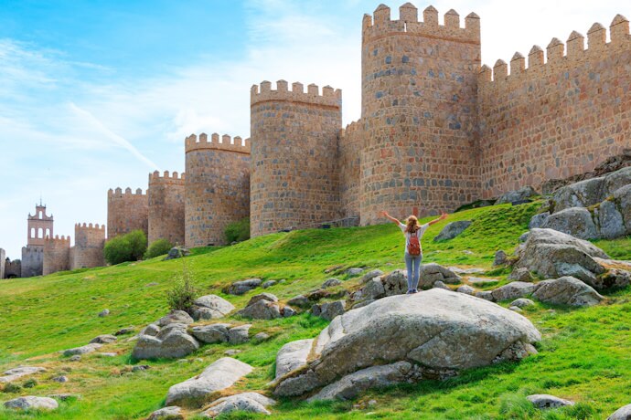 A woman with arms raised stands on rocks in front of the medieval stone walls and towers of Ávila, Spain, symbolizing exploration and heritage.