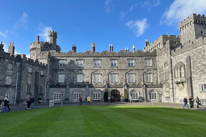 Wide view of Kilkenny Castle’s gray stone façade and towers framing a central courtyard, with a bright green lawn in the foreground and a clear blue sky overhead.