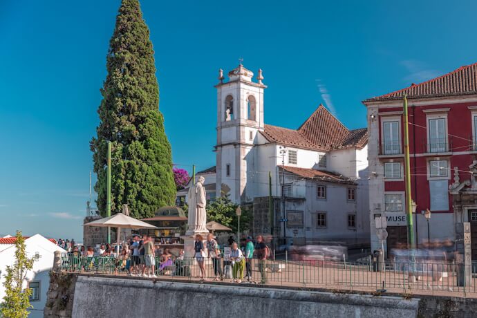 A lively terrace in Lisbon with people gathered near a church the St Vincente statue, enjoying the sunny view over the city.