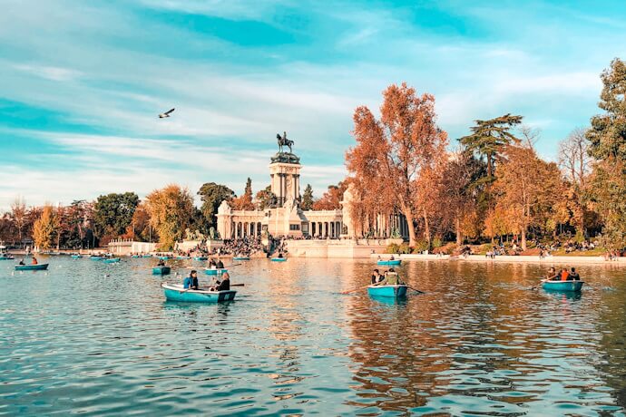 Rowboats drifting on a calm lake in Retiro Park, with the grand Alfonso XII monument framed by autumn-colored trees.