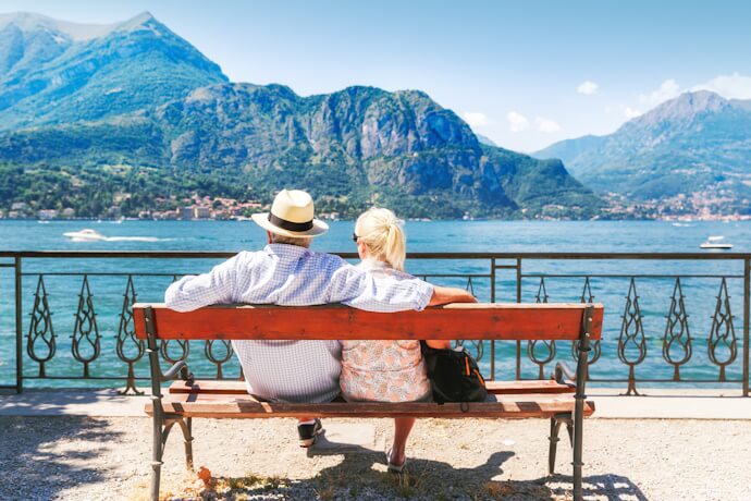 A senior couple sitting on a bench overlooking the serene waters of Lake Como, with dramatic green mountains rising in the background under a bright blue sky.