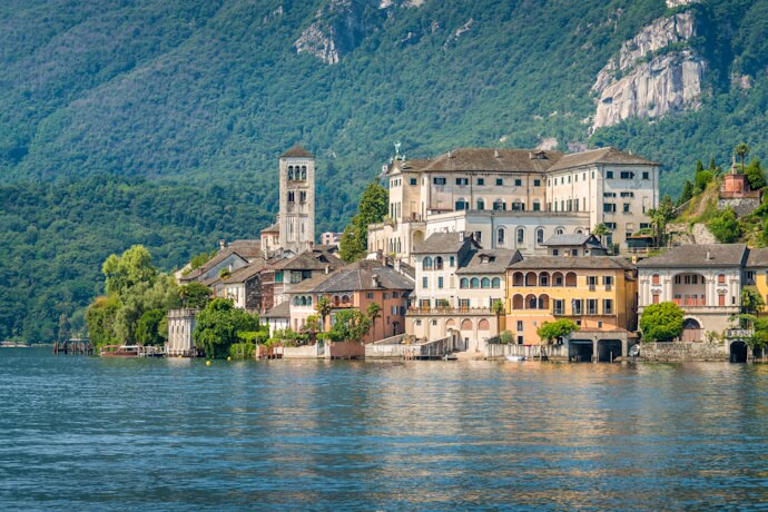 Charming lakeside village of Orta San Giulio on Lake Orta, with pastel-colored buildings, a church bell tower, and forested hills rising behind it.