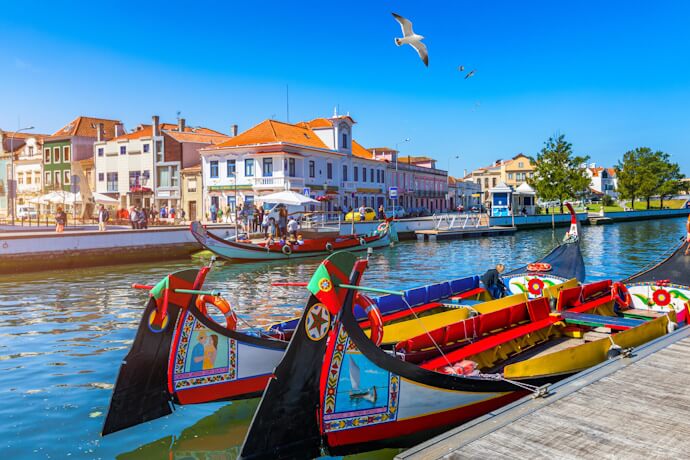 Traditional moliceiro boats with colorful painted prows floating on the calm canal waters of Aveiro, surrounded by pastel-hued buildings under a bright blue sky.