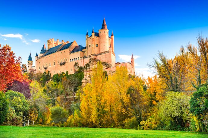 The fairytale-like medieval castle of Segovia, Spain, standing on a rocky hilltop surrounded by autumn-colored trees.