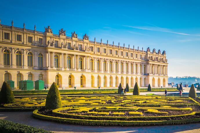 The elegant façade of the Palace of Versailles with its ornate windows, fronted by manicured geometric gardens in the bright morning light.