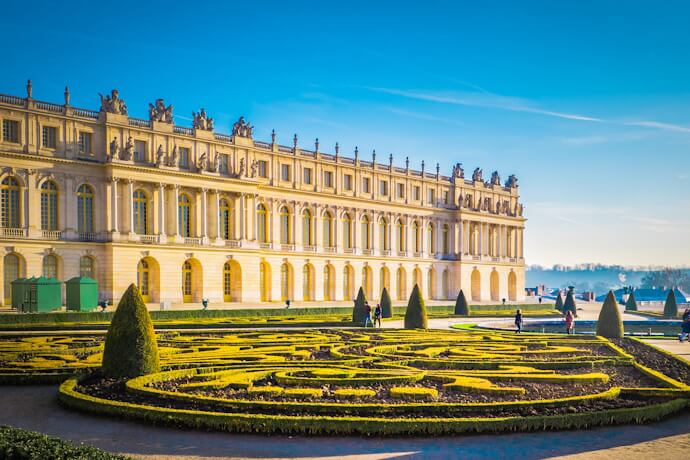 View of the Palace of Versailles and its manicured gardens featuring geometric hedges and sculptures under a clear blue sky.