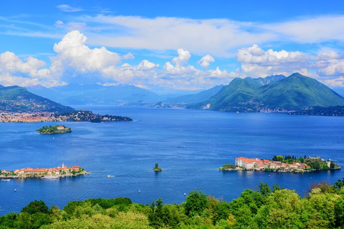A stunning panoramic view of Lake Maggiore in northern Italy, showcasing the lush Borromean Islands scattered across the deep blue water, with forested hills and the Alpine mountains in the background under a bright, cloud-dappled sky.