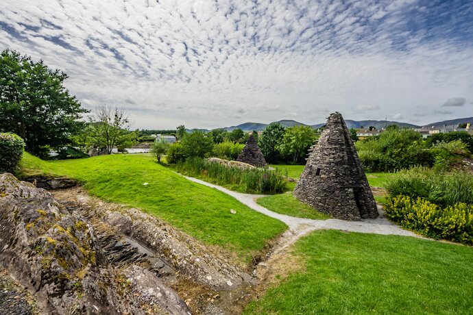 Stone beehive huts sit beside winding gravel paths in a lush green field near Sneem, with trees and distant hills under a dramatic, cloud-streaked sky.