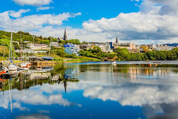 Panoramic view of Galway’s riverside with colorful houses and church spires on a green hillside, small boats moored along the shore, and calm water reflecting the blue sky.