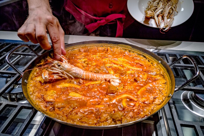 Close-up of a seafood paella simmering in a pan, as a cook places a large prawn on top.