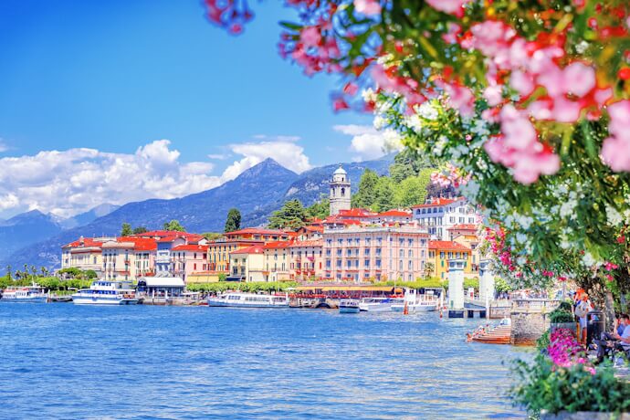 Picturesque view of the town of Bellagio on Lake Como, with flower-adorned promenade, pastel buildings, and boats moored along the waterfront against a backdrop of mountains.