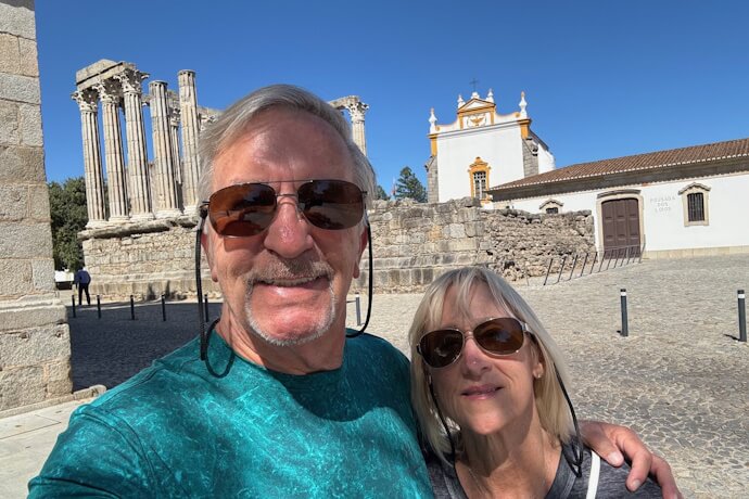 A couple smiling in front of the Roman Temple of Évora and nearby historic buildings on a clear blue day.