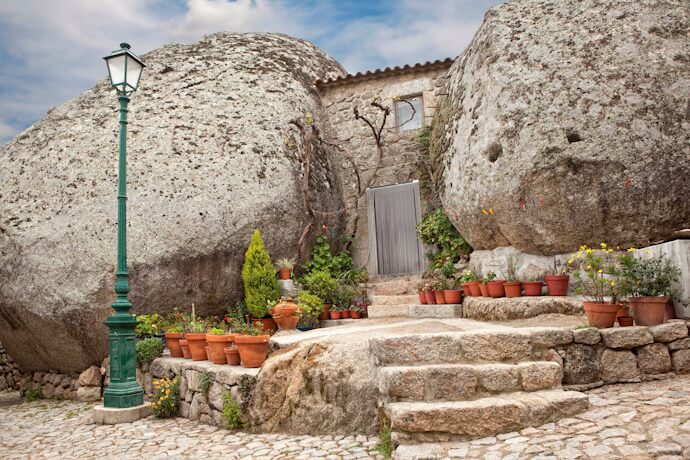A scenic view of Monsanto, Portugal, known as "the most Portuguese village". The image showcases stone houses with red-tiled roofs built among massive boulders, including one large rock resting on top of a house.