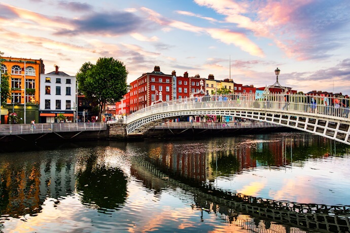 The white Ha’penny Bridge arches over the River Liffey in Dublin at sunset, with colorful riverside buildings behind it and the bridge reflected in the calm water below.