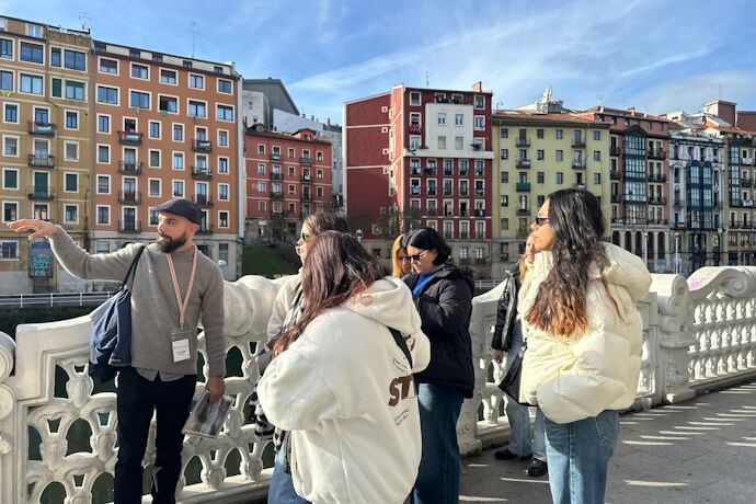Guided walking tour in Bilbao with a local guide explaining the city’s history to the team gathered on a bridge with colorful riverside buildings in the background.