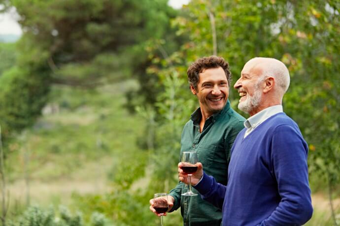 Two men smiling and clinking glasses of red wine outdoors, surrounded by green vineyard scenery.