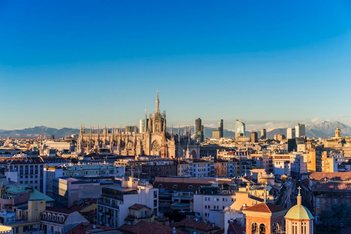 A panoramic view of Milan at sunset, with the iconic Milan Cathedral (Duomo di Milano) rising above the city skyline and the Alps in the distant background.