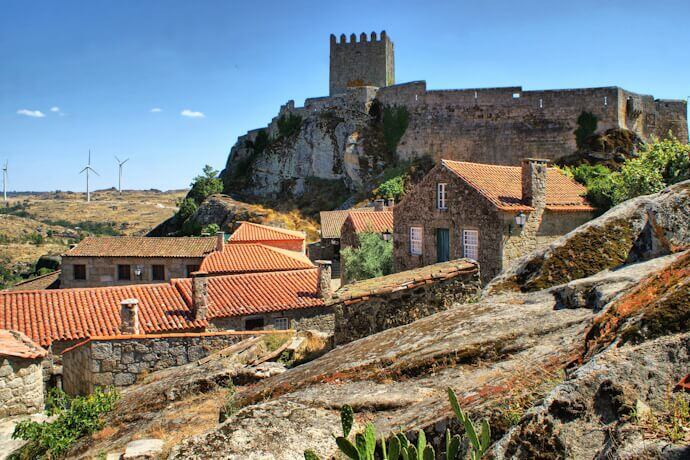 A picturesque view of Sortelha, featuring stone houses with red-tiled roofs nestled among rocky terrain. In the background, a medieval castle with a crenelated tower stands atop a hill, overlooking the village.