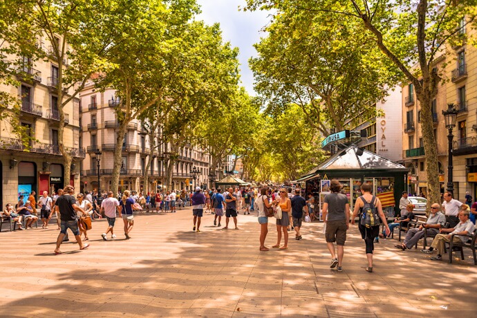 Bustling Barcelona promenade shaded by tall trees, with pedestrians, kiosks, and café seating along the wide walkway.