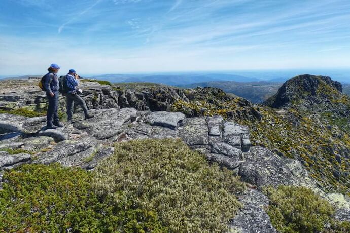 Adventurous couple standing on a rocky outcrop in Serra da Estrela Natural Park, with a view of mountains and a blue sky in the background.