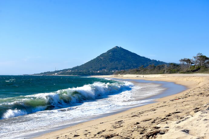 Sandy beach with waves crashing against the shore, with Monte de Santa Tecla rising in the background.