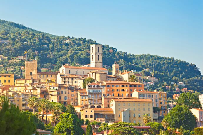 Panoramic view of the hilltop town of Grasse, France, with pastel buildings, a church tower, and lush green hills in the background.