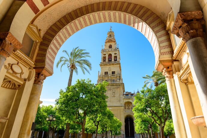 View through Moorish arches of the Great Mosque–Cathedral of Córdoba, with palm trees, ornate columns, and the bell tower centered in the frame.