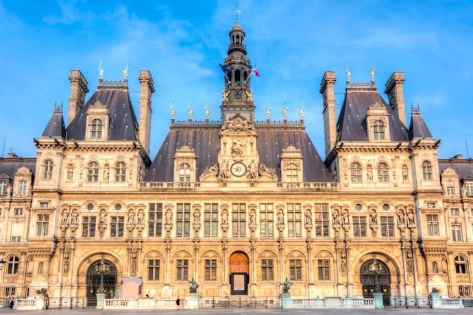 The ornate façade of Hôtel de Ville in Paris glowing in soft morning light against a blue sky.