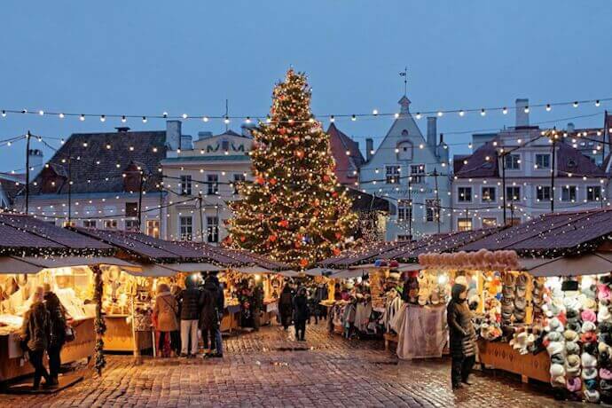 A festive market square in Paris at twilight with a towering Christmas tree, wooden stalls decorated with lights, and shoppers browsing.