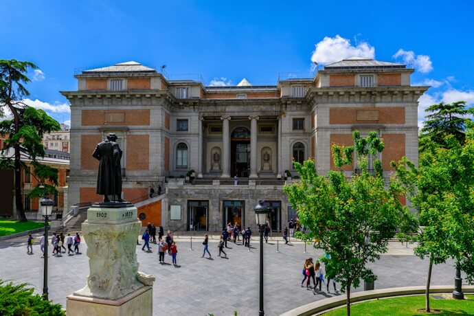 Exterior of the Prado Museum in Madrid, Spain, with people walking in the plaza and a statue of painter Diego Velázquez in the foreground.