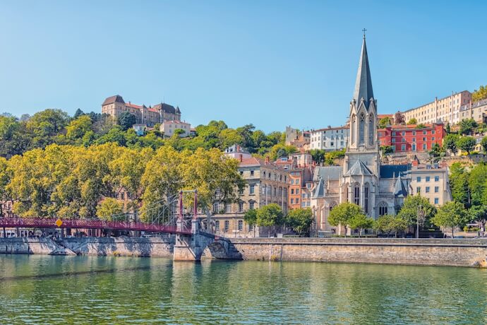 Picturesque view of the Saône River in Lyon with colorful buildings, a church spire, and a red pedestrian bridge surrounded by greenery.