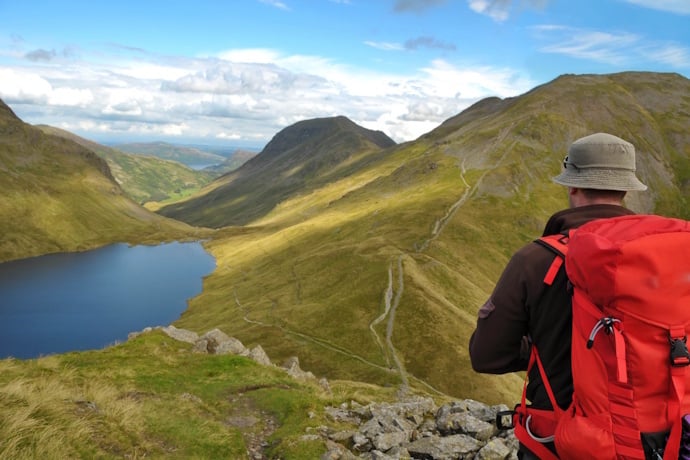 Hiker with a red backpack admiring the vast mountain scenery and blue lake below in the Lake District.