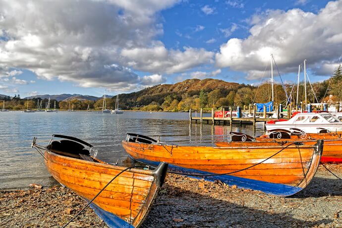 Wooden rowing boats resting on the shore of Windermere Lake surrounded by autumn-colored hills and blue skies.