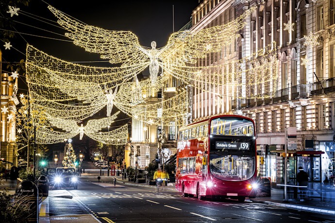A classic red double-decker bus driving under dazzling golden angel-shaped Christmas lights along Regent Street in London.