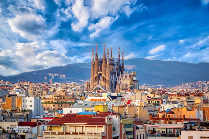 Barcelona skyline featuring the Sagrada Família’s soaring spires rising above colorful city rooftops under a dramatic blue sky.