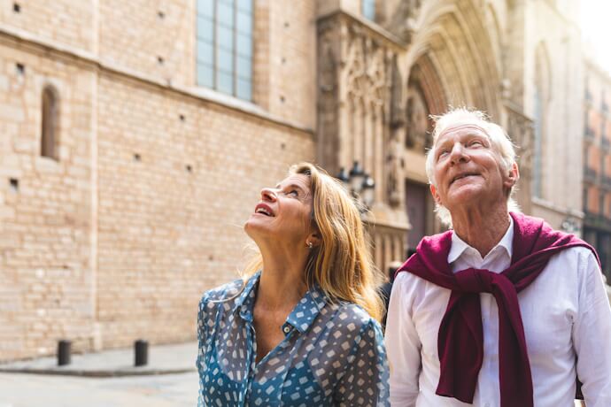 Two senior travelers stroll beside a historic stone cathedral, looking up at the architecture in warm afternoon light.