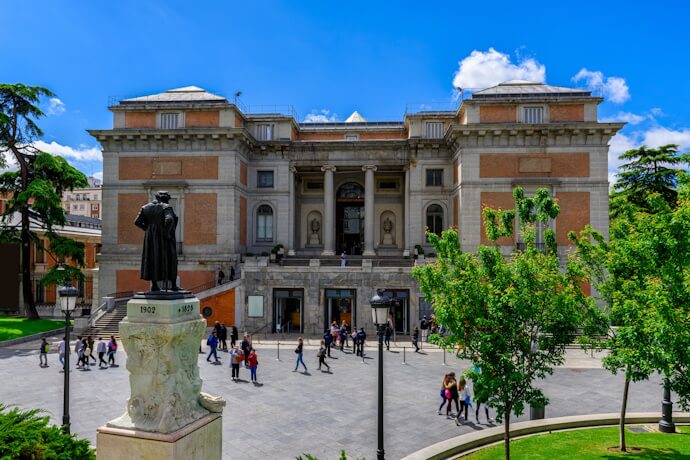 Exterior of the Prado Museum in Madrid with visitors walking across the plaza and a statue of Spanish painter Velázquez in the foreground.