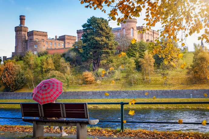 A peaceful riverside moment beneath falling leaves, with a pop of color from a red umbrella and Inverness Castle watching from above.