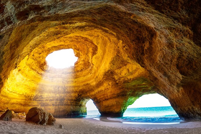Inside Benagil Cave in the Algarve, showing golden rock formations, a natural skylight opening above, and sunlight illuminating the sandy beach and ocean entrance