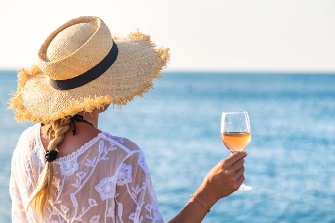 Woman wearing a straw hat and sheer white cover-up, holding a glass of rosé wine while looking out over the calm blue sea on a sunny day.