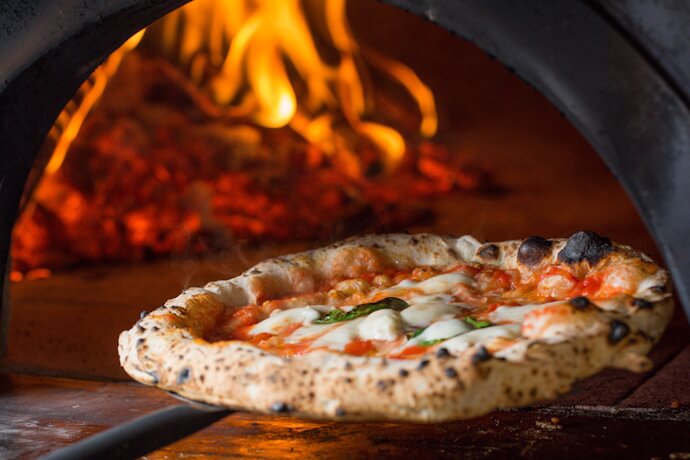 Neapolitan pizza baking in a traditional wood-fired oven, with flames glowing in the background.