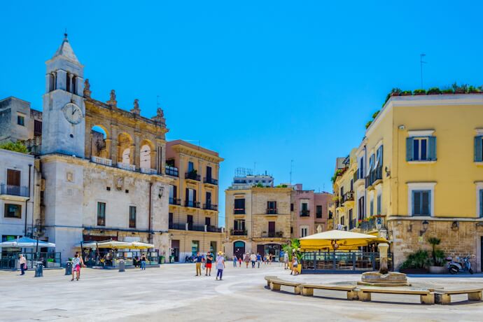 Piazza scene in Bari’s old town with historic buildings, a church façade, and people walking across the square.