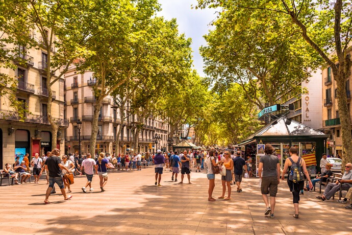 Crowds stroll along Barcelona’s tree-lined La Rambla, surrounded by historic buildings and shaded kiosks.