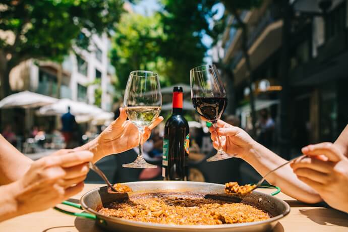 Close-up of a shared outdoor meal with paella, wine glasses, and a bottle, on a sunny Barcelona street café.